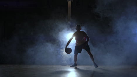 A-man-with-a-basketball-on-a-dark-basketball-court-against-the-backdrop-of-a-basketball-ring-in-the-smoke-shows-dribbling-skills-illuminated-by-three-lanterns-in-backlight